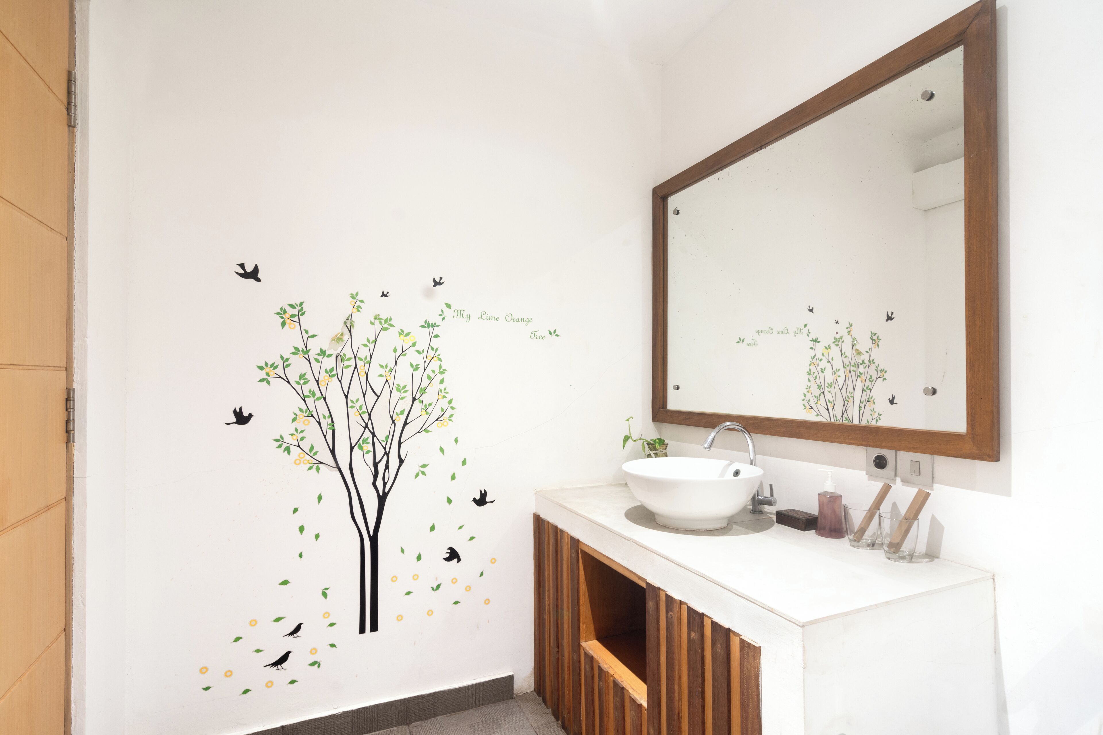 Un baño con lavamanos blanco, tocador de madera y un adhesivo mural con un árbol y pájaros.