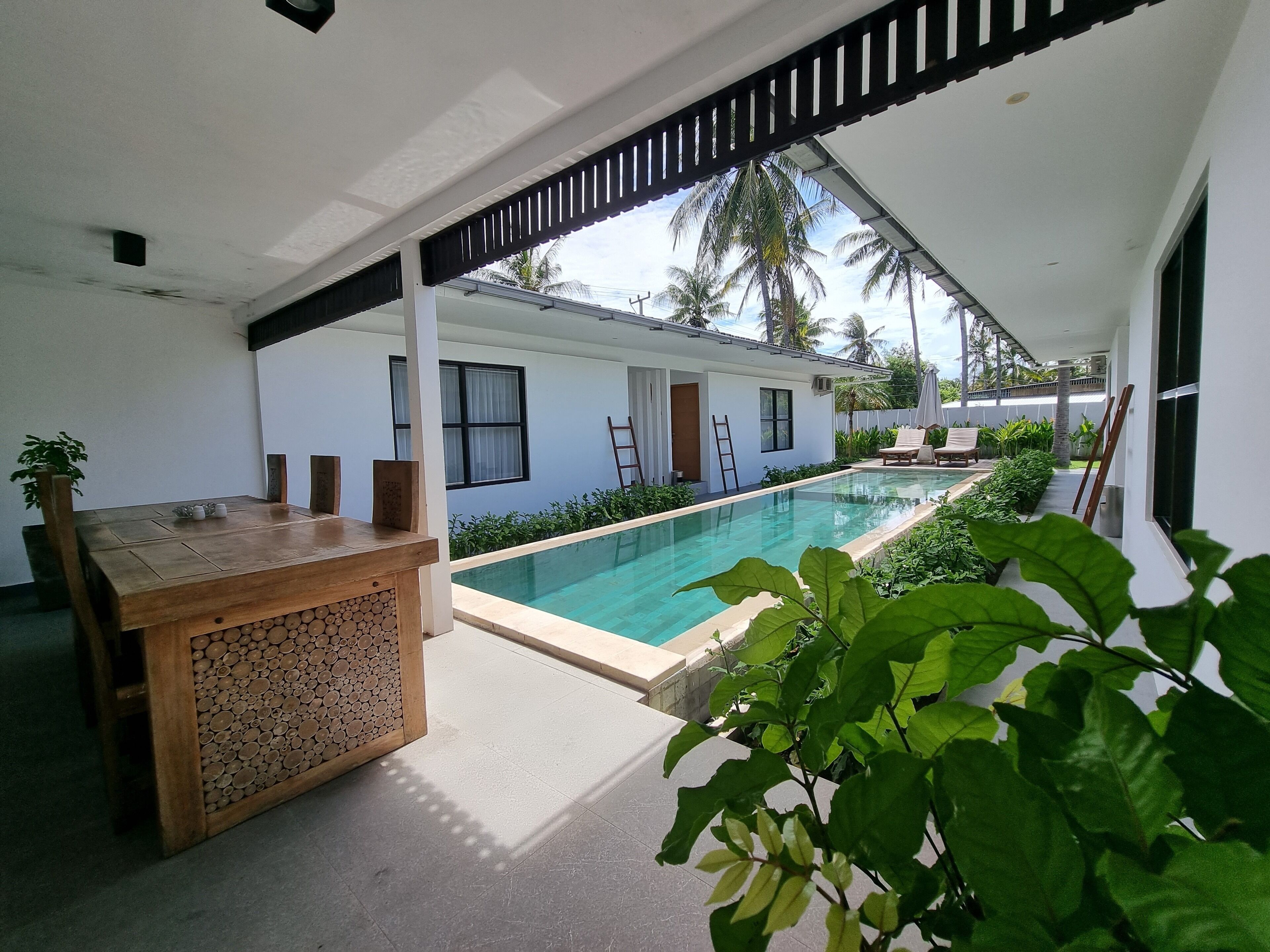 A poolside area with a wooden table and chairs, a white building with windows, and a view of palm trees.