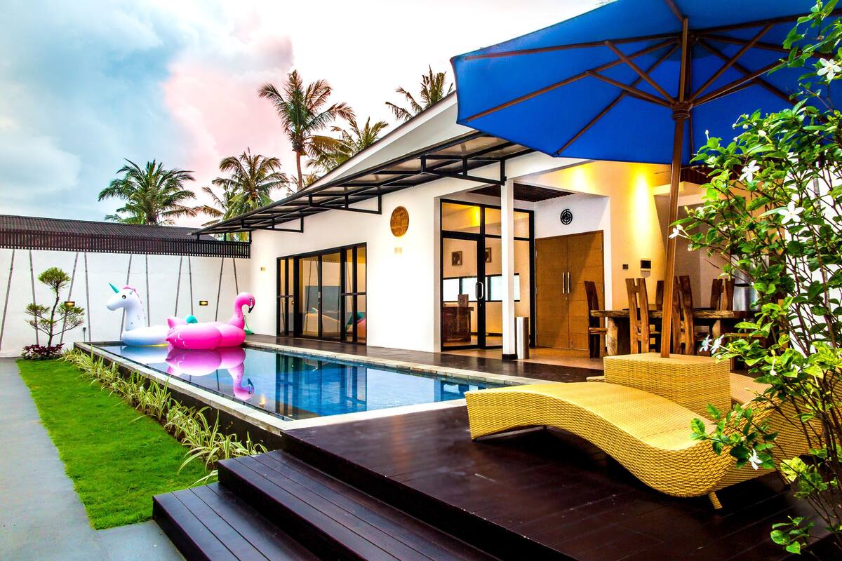 A modern outdoor pool area with a lounge chair, a floating unicorn decoration, and a blue umbrella.