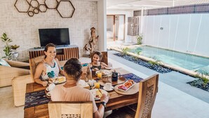A dining area with a pool view, wooden furniture, and a TV in the background.