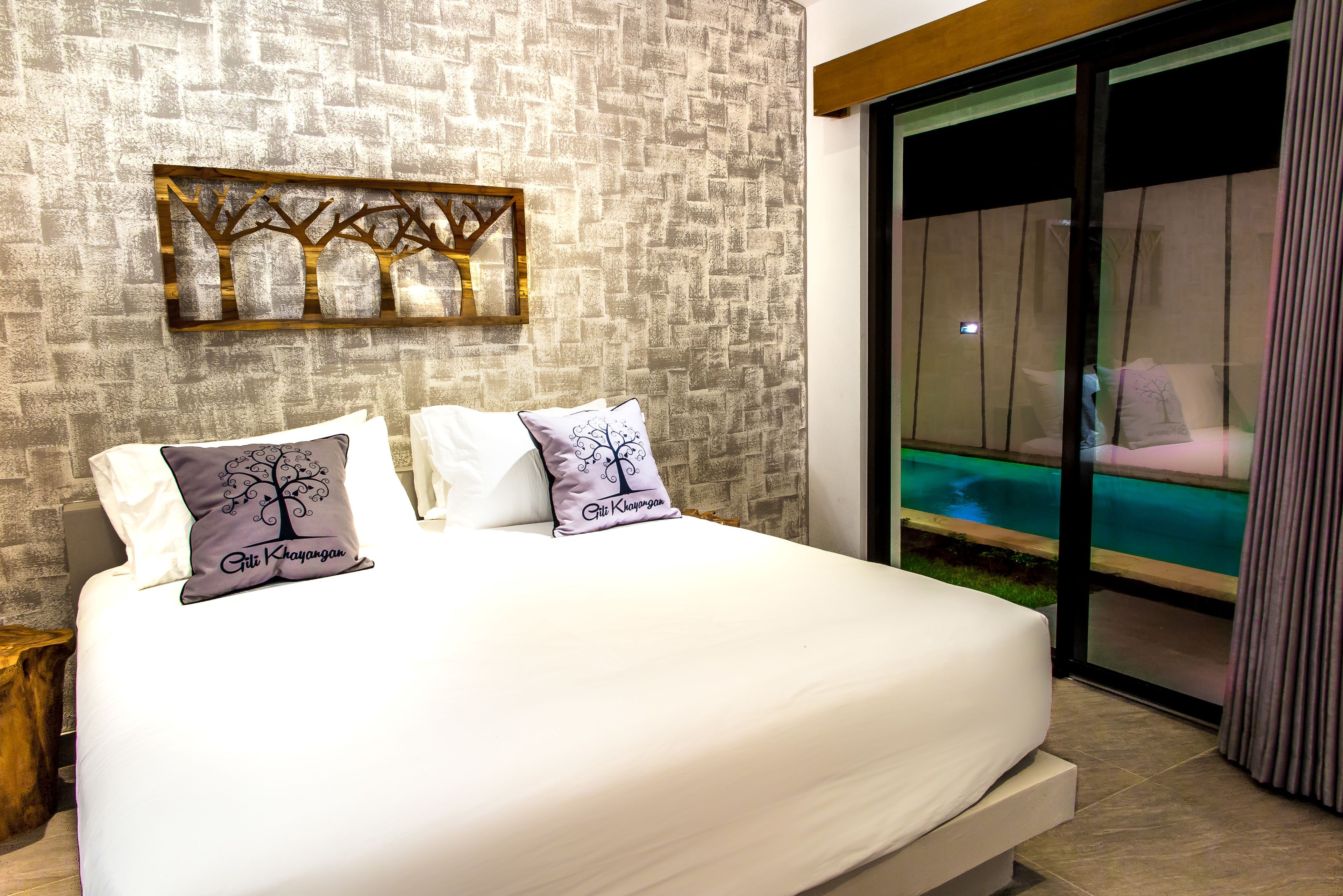 A bedroom with a large bed, two pillows with tree designs, a wall-mounted artwork, and a view of a pool through a glass door.
