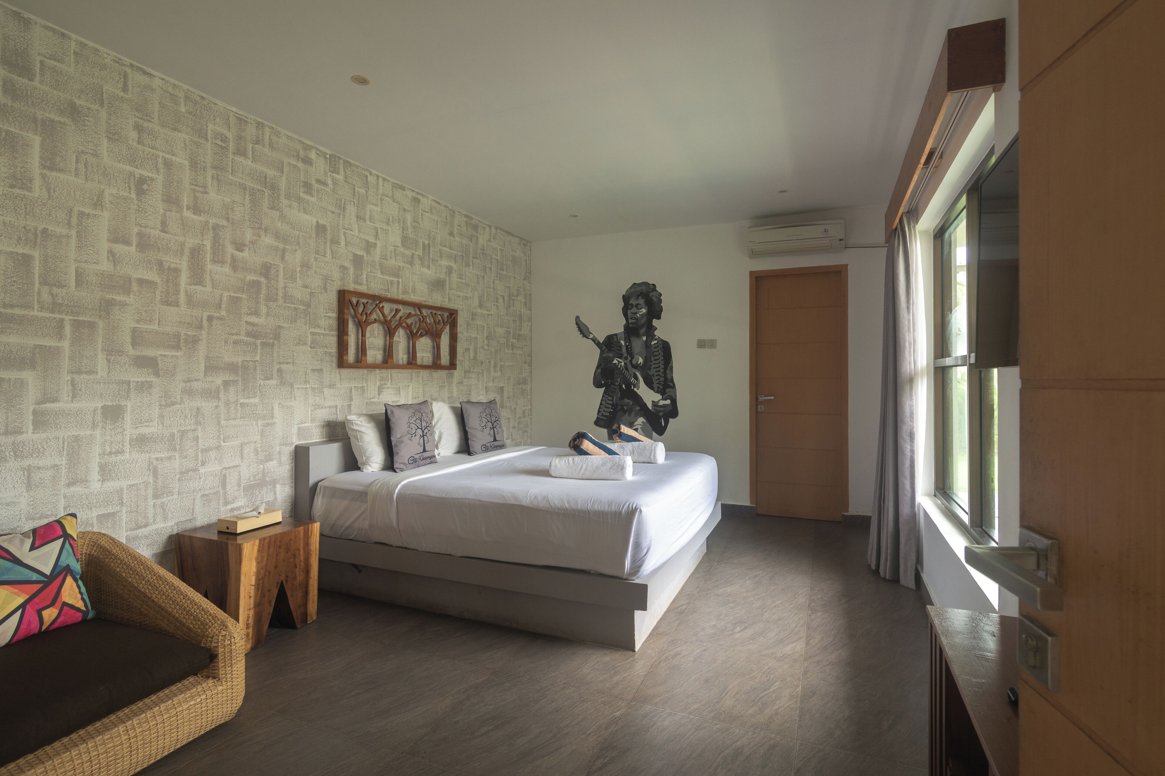 A modern hotel room with a large bed, a wooden headboard, a small wooden side table, a wicker sofa, and a large window with curtains.