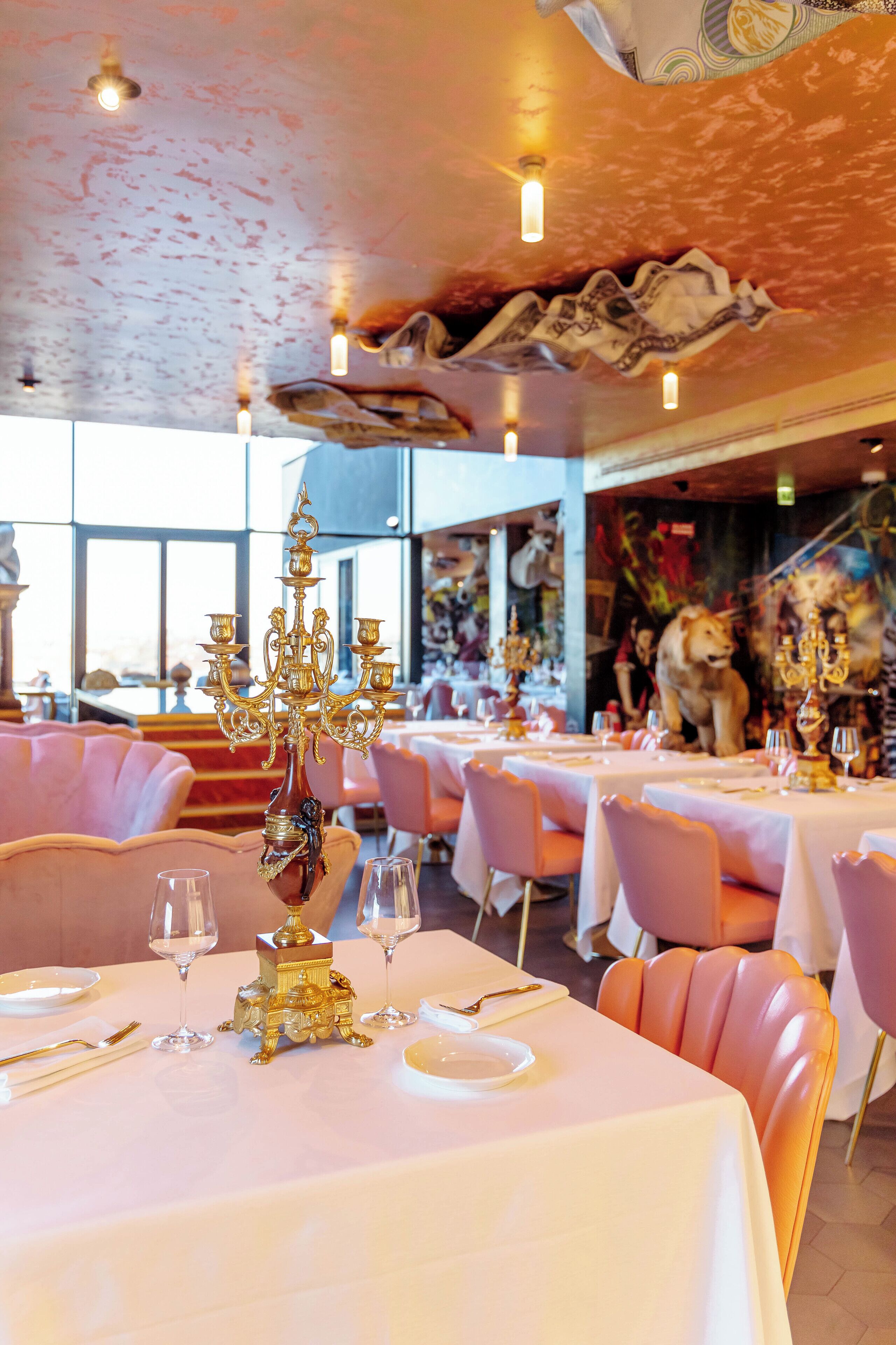 A restaurant with tables set for dining, featuring gold candelabras and pink chairs.