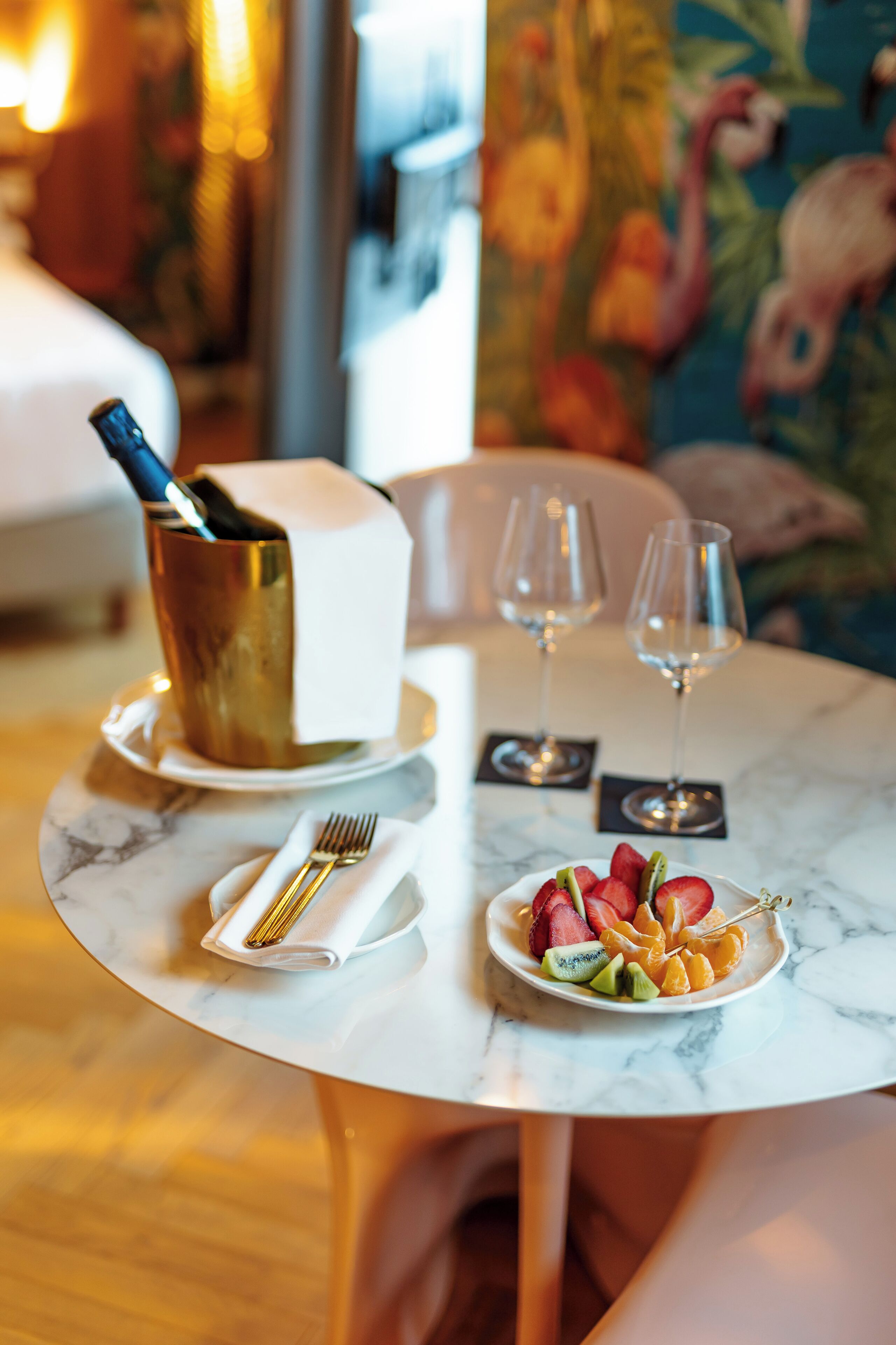 A dining table with a white plate of fruit, a gold-colored bottle, and two wine glasses.