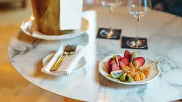 A dining table with a white plate of fruit, a gold-colored bottle, and two wine glasses.