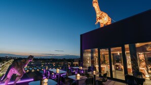 Een restaurant op het dak met tafels en stoelen, een girafbeeld en uitzicht over de stad.