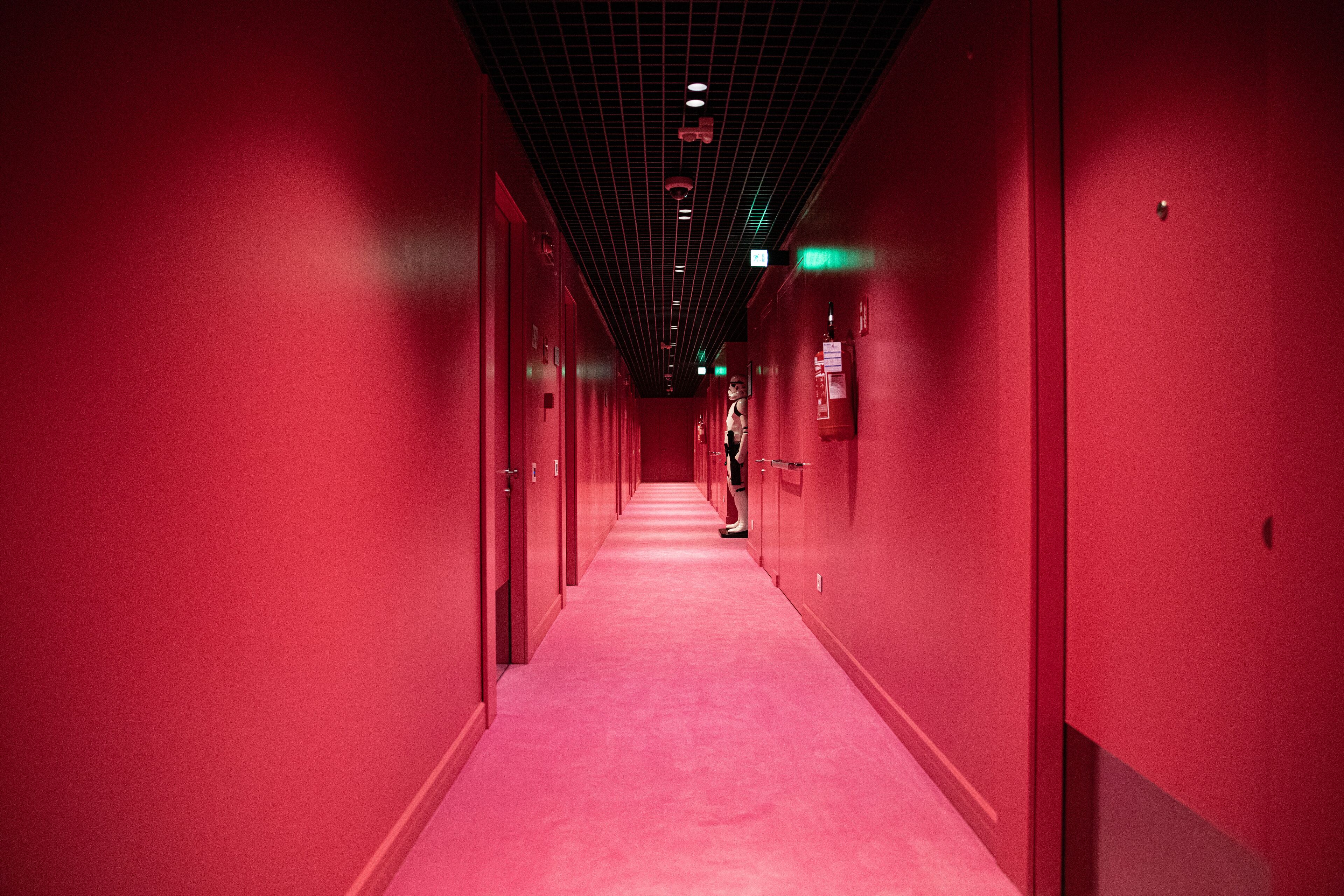 A long, narrow corridor with red walls and a red carpet, leading to a door at the end.