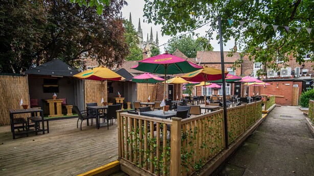 An outdoor patio with wooden tables and chairs, surrounded by a wooden fence with climbing plants.