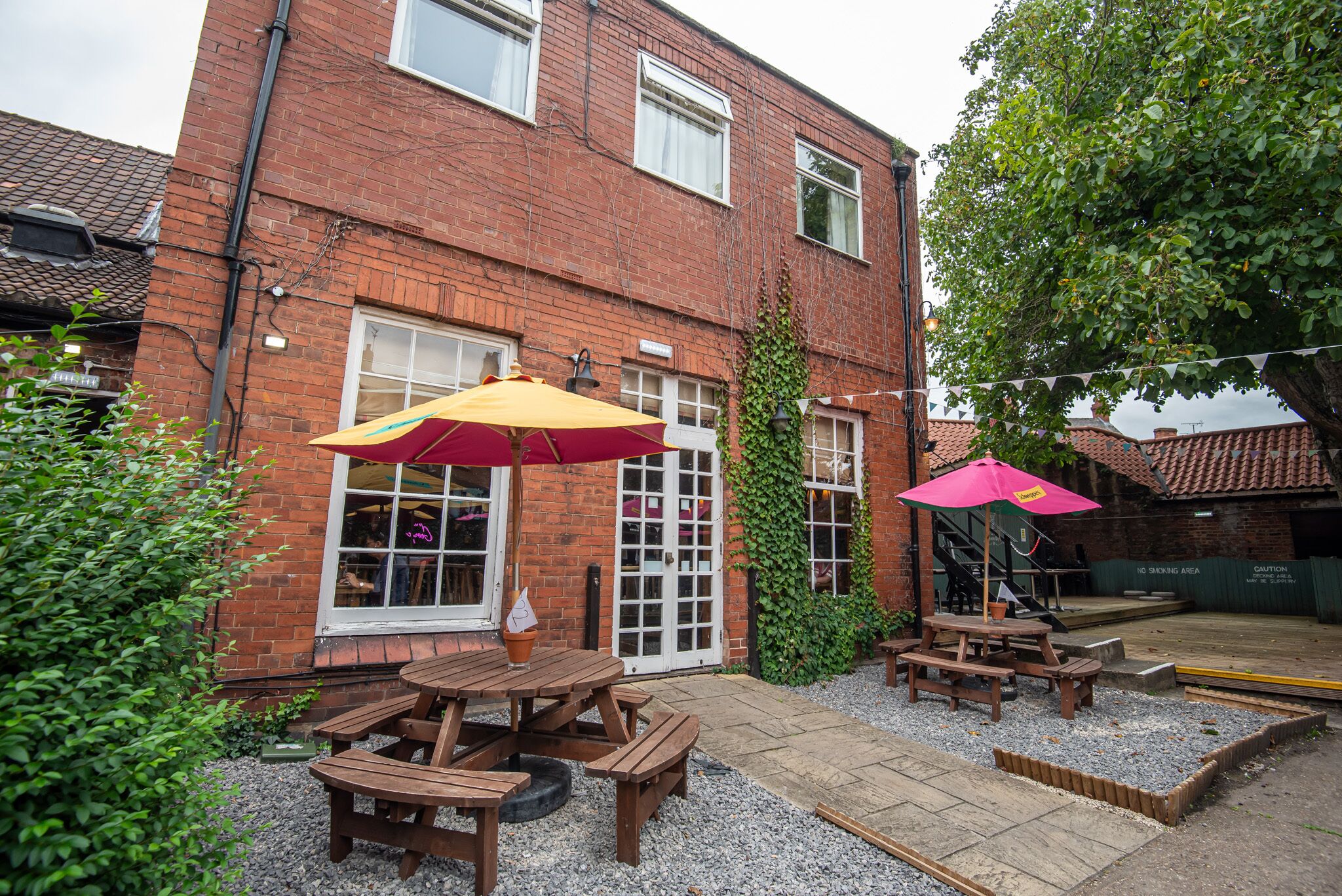 A brick building with a patio area featuring wooden picnic tables and two umbrellas.