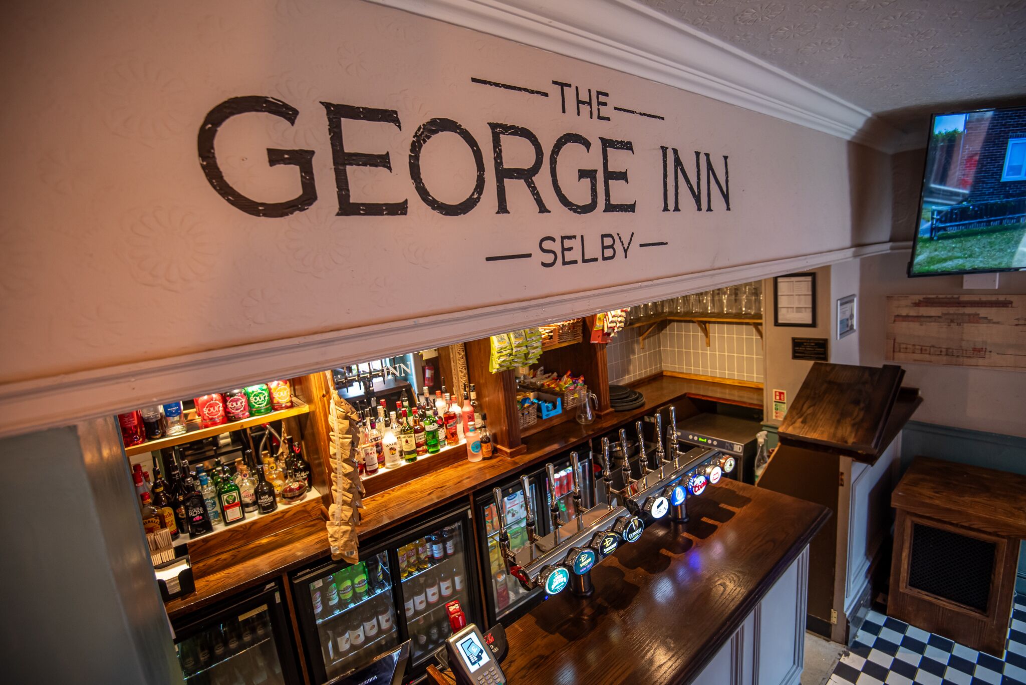 A bar area with a sign reading 'The George Inn Selby,' a selection of bottled beverages, and a television mounted on the wall.