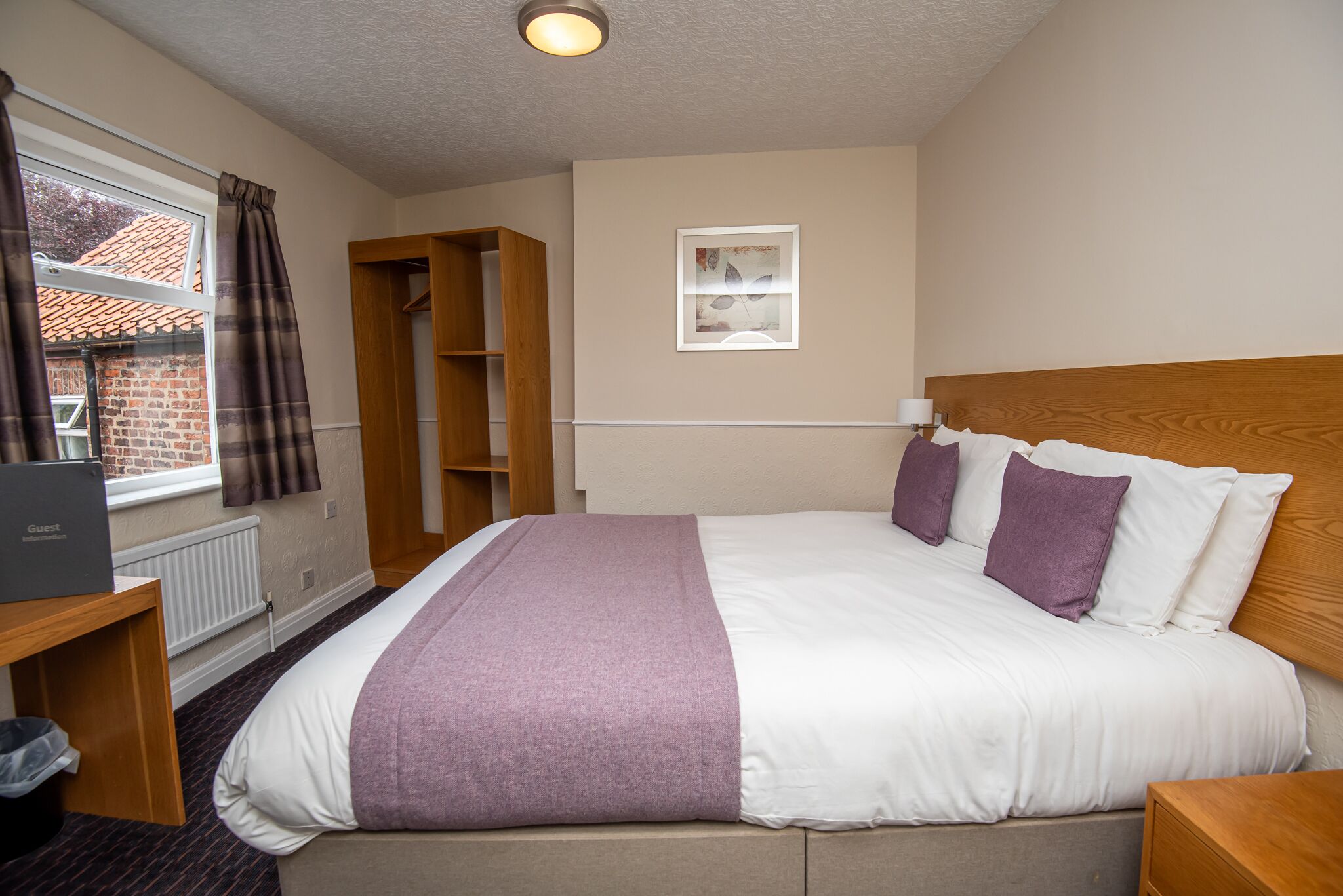 A hotel room with a bed, a desk with a laptop, a window with curtains, a wooden wardrobe, and a small picture on the wall.