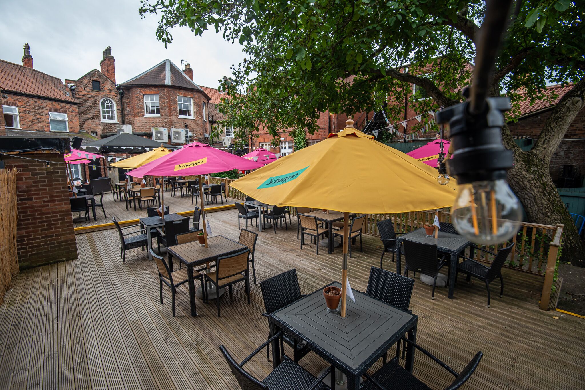 An outdoor patio with tables and chairs, umbrellas, and brick buildings in the background.