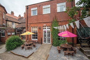 A brick building with a patio area featuring wooden tables and benches, a small garden, and a white door. - The George Inn (Selby)