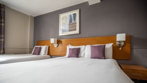 A hotel room with two beds, a wooden headboard, wall-mounted nightstands, a bedside table, and a framed artwork on the wall.