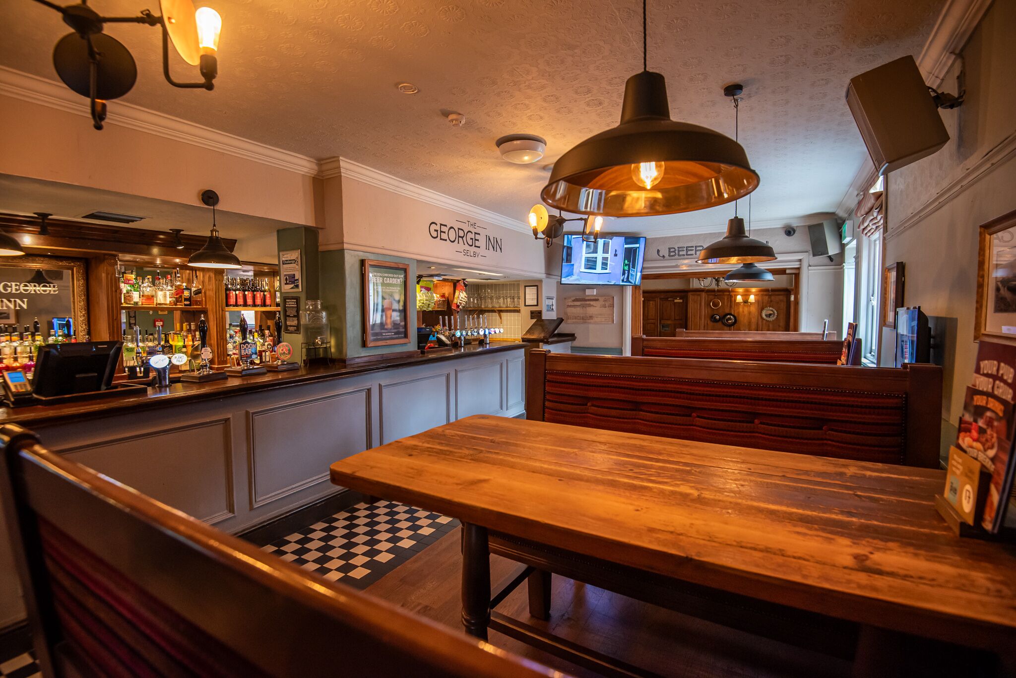 A bar area with wooden benches, a checkered floor, and a bar counter with various bottles and a cash register.