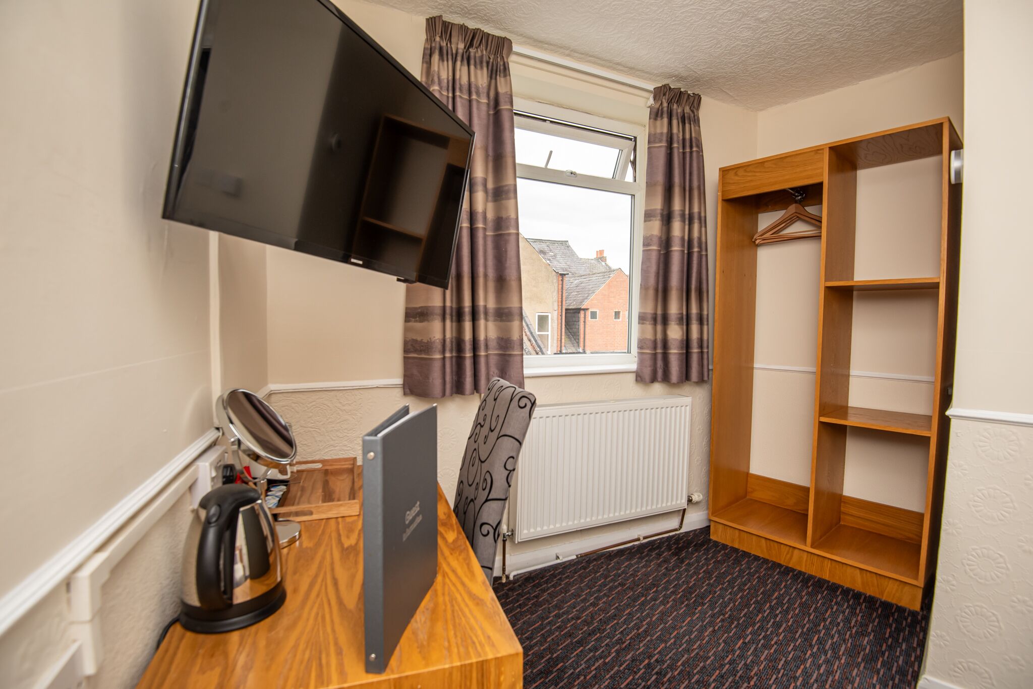 Quarto de hotel com escrivaninha, uma cadeira, televisão fixada na parede, radiador, janela com cortinas e guarda-roupa de madeira.