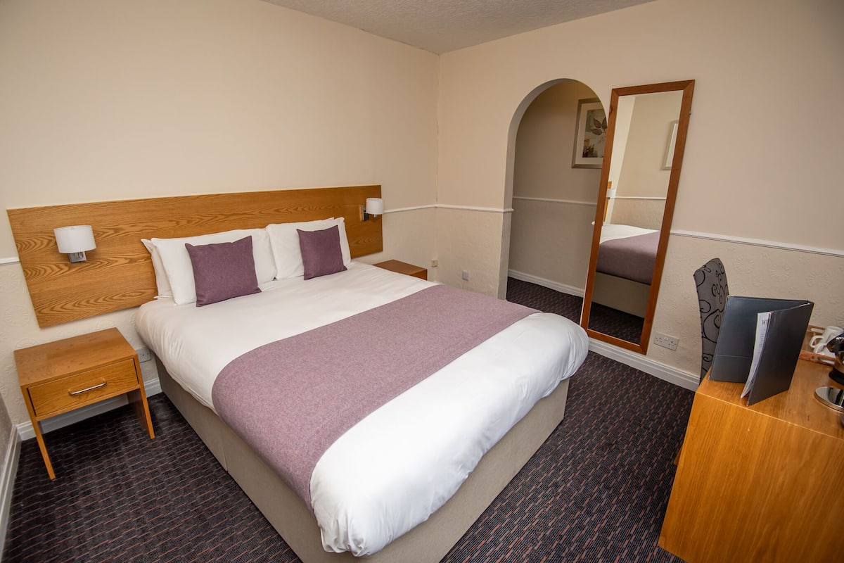 A hotel room with a bed, nightstand, mirror, and a desk with a computer.