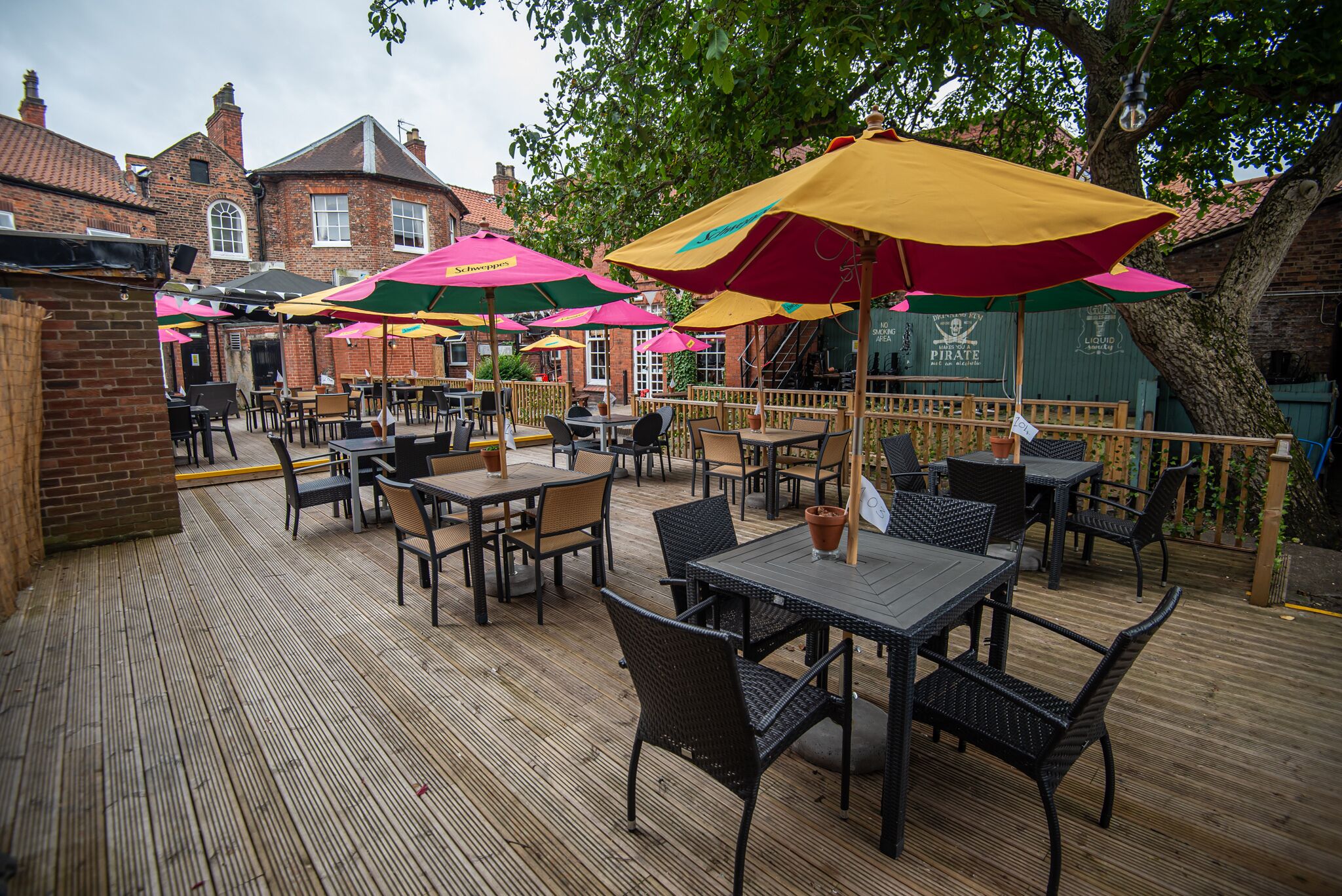 An outdoor patio with tables and chairs, umbrellas, and a brick building in the background.