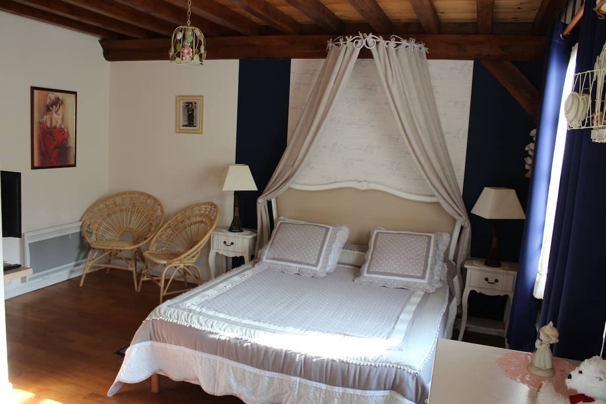 A bedroom with a canopy bed, two wicker chairs, bedside tables with lamps, and a decorative birdcage.