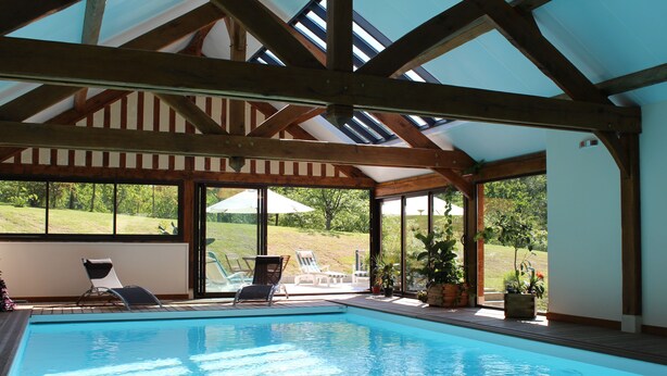Piscina interna com vigas de madeira, grandes janelas e cadeiras de descanso.