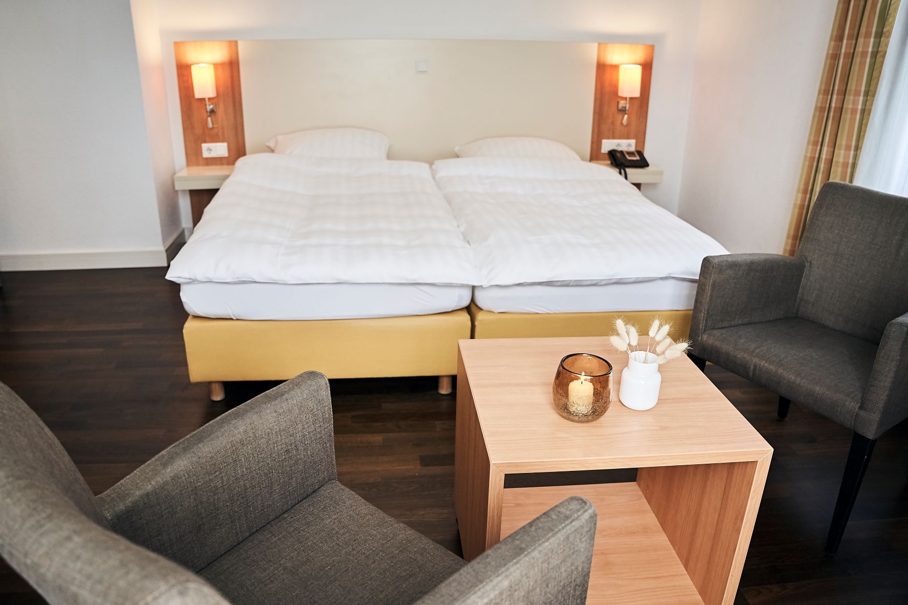 A hotel room with a double bed, bedside lamps, a small table with a candle and decorative items, and two armchairs.