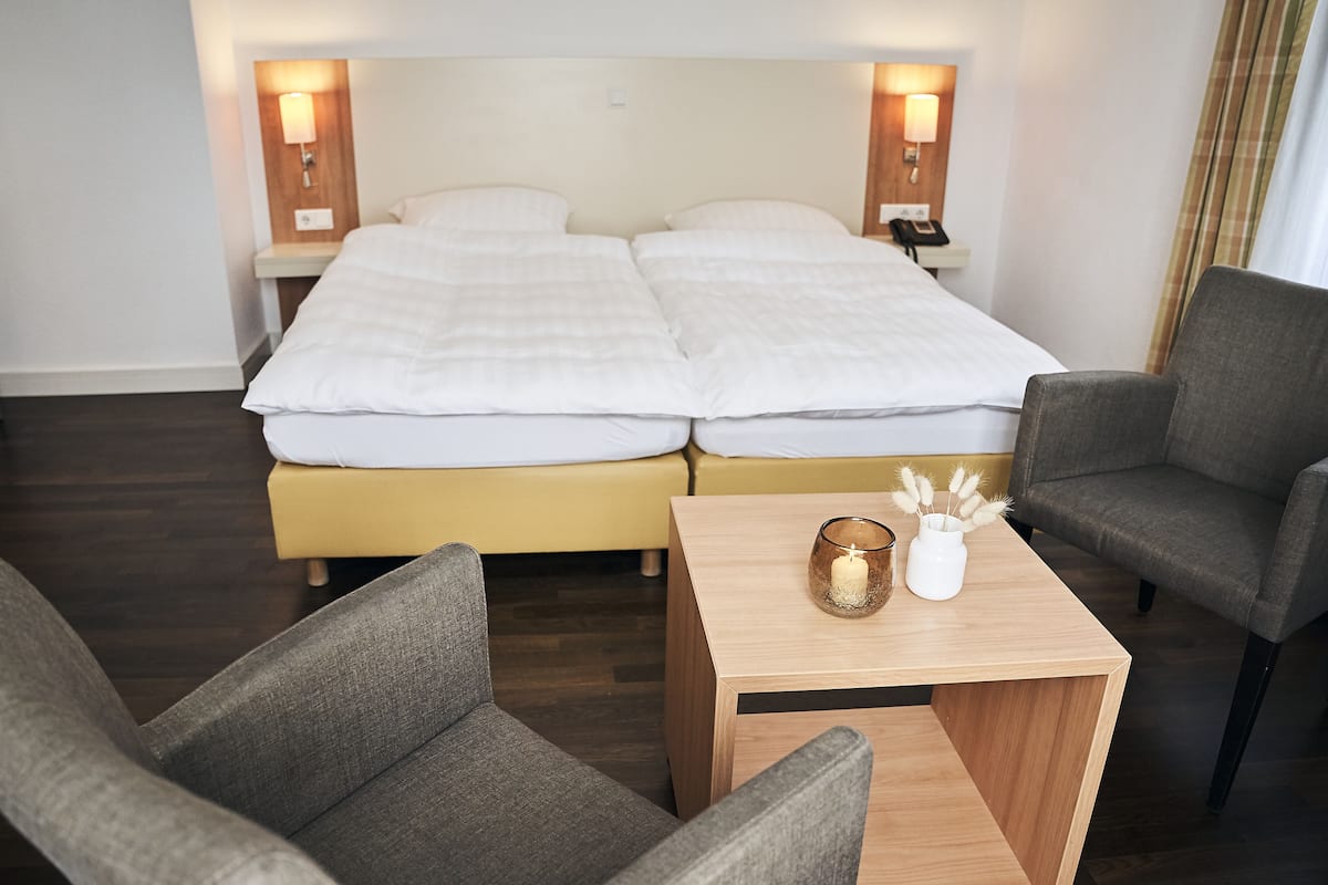 Habitación de hotel con una cama doble, dos mesitas de noche con lámparas, una pequeña mesita de madera con una vela y objetos decorativos, y dos sillones grises.