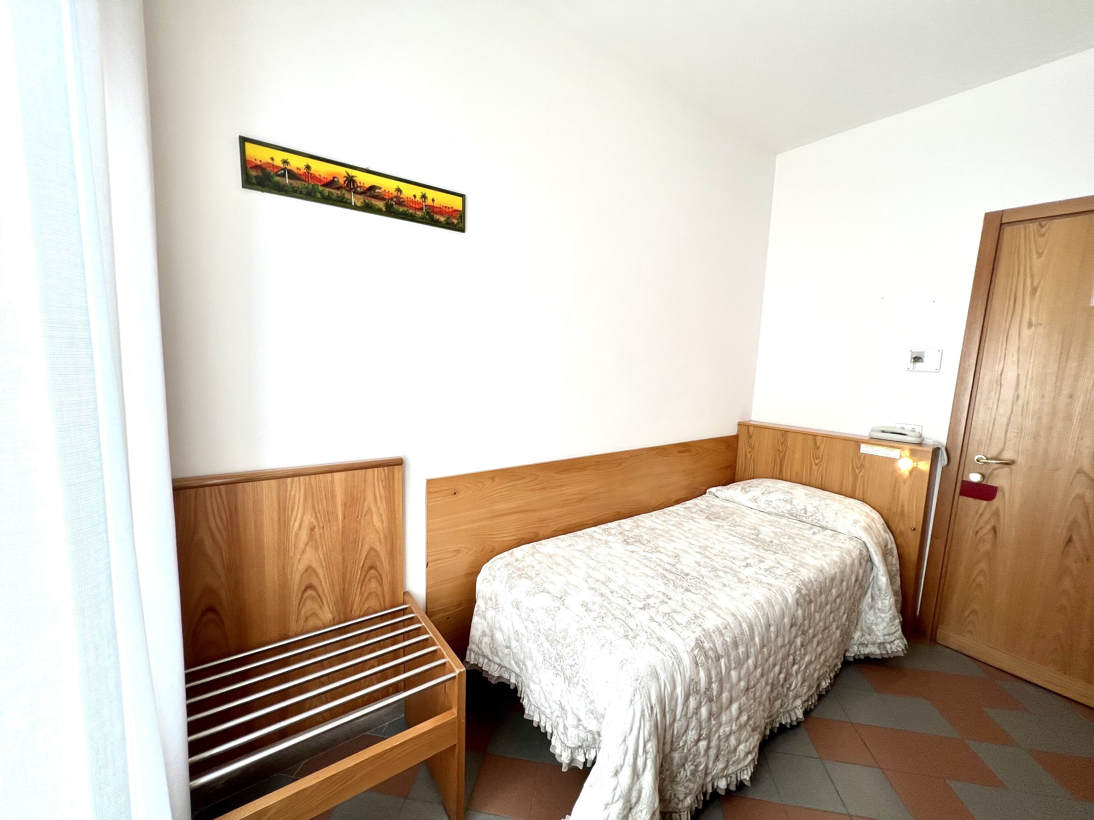 Una stanza piccola e ordinata con un letto singolo, una panca di legno e una porta.