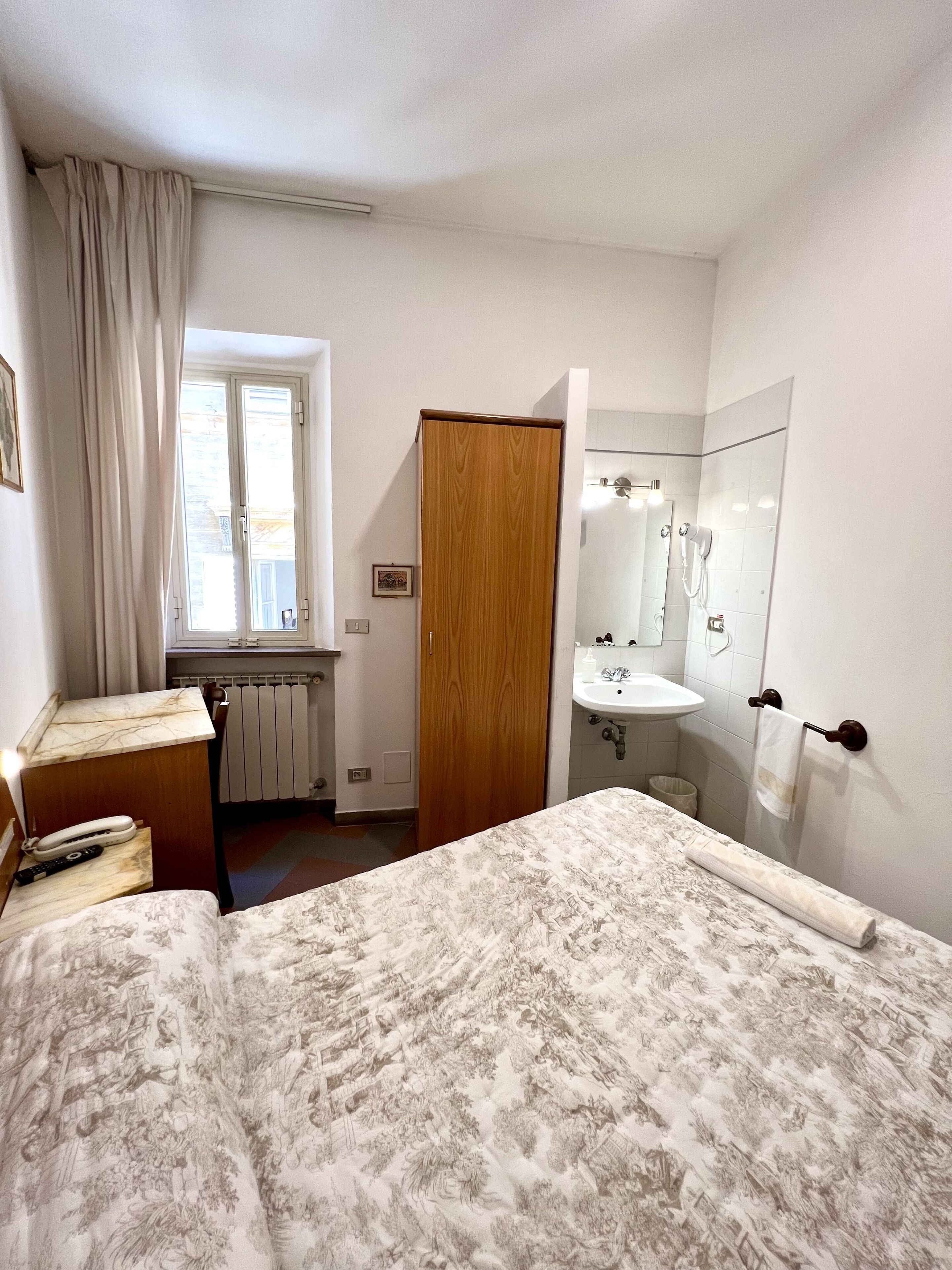 Une chambre à coucher avec un lit, une armoire en bois, une salle de bain avec un lavabo et un miroir, et une fenêtre avec des rideaux.