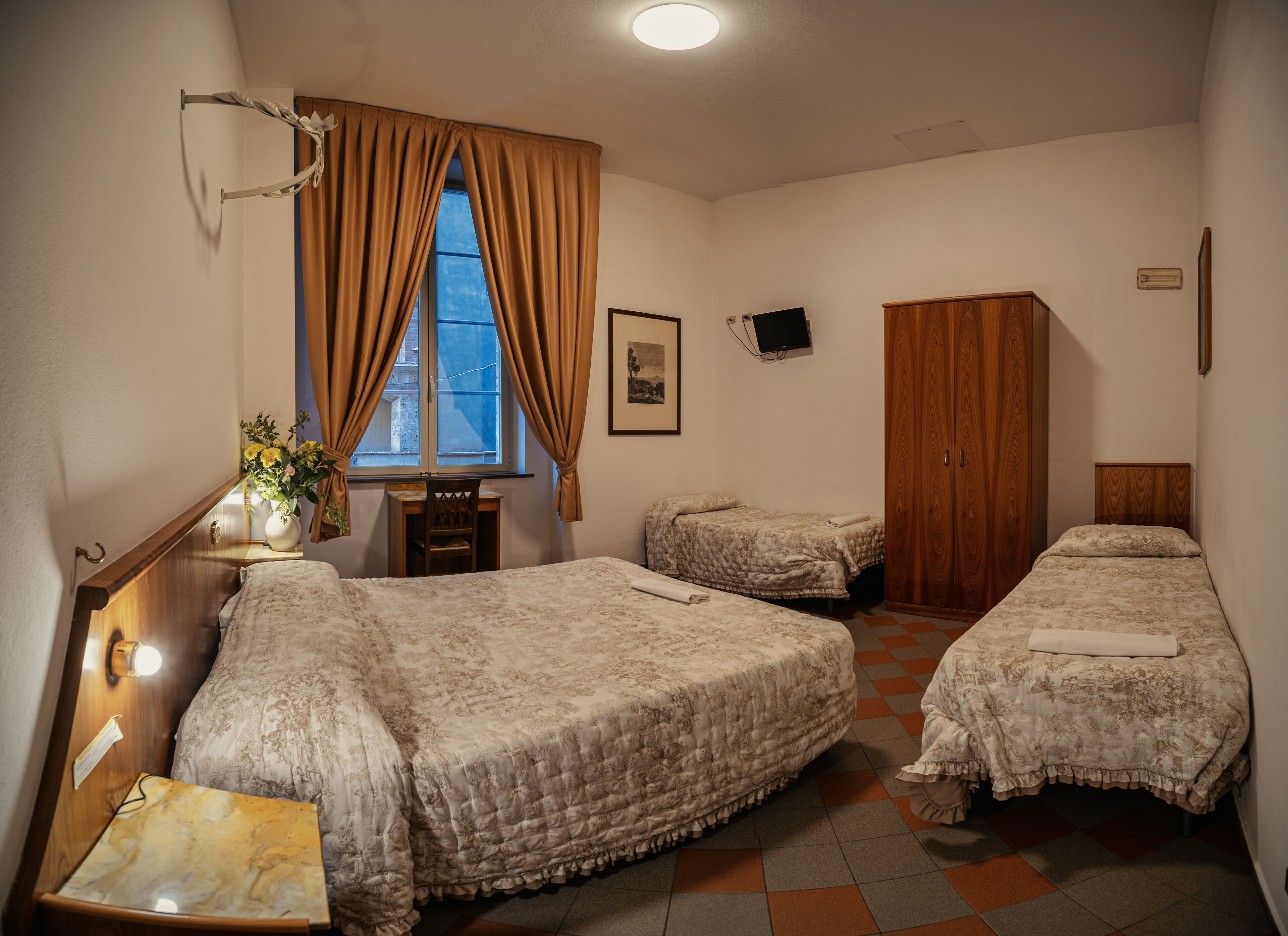A hotel room with two beds, a desk, a chair, a window with curtains, a wooden wardrobe, and a wall-mounted TV.