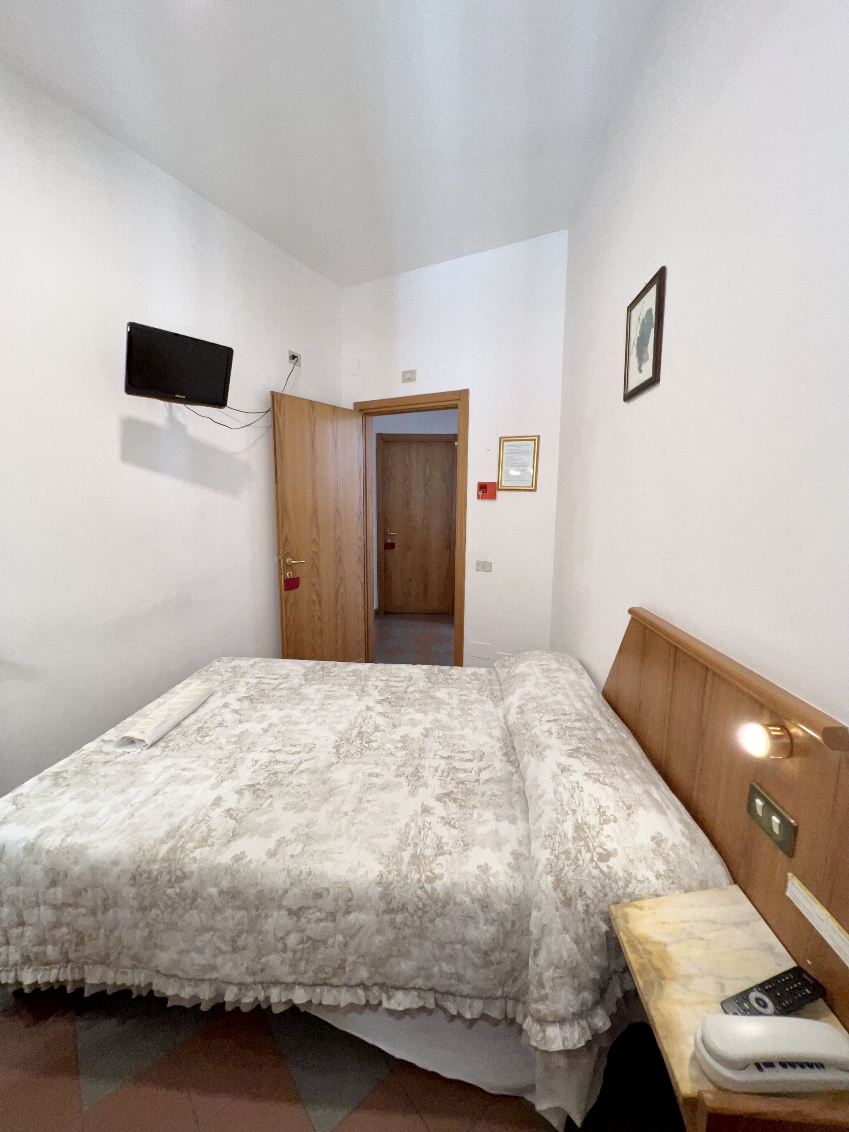 Ein Schlafzimmer mit Bett, Nachttisch, Wand-Fernseher und einer Tür zu einem anderen Raum.