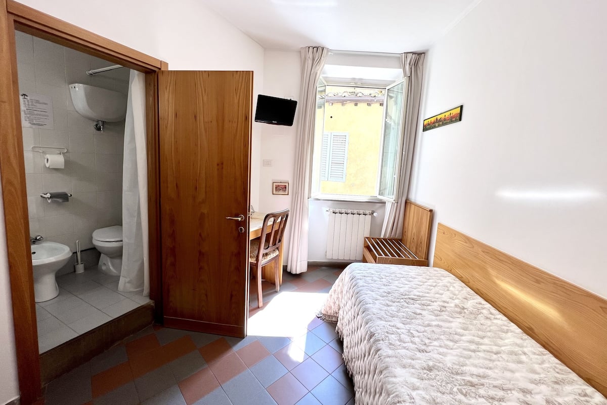 A bedroom with a bed, a wooden door leading to a bathroom, a window with curtains, and a radiator.