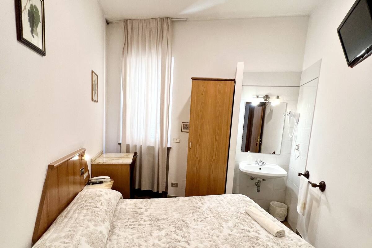 A bedroom with a bed, a wooden wardrobe, a bedside table, a mirror, and a television mounted on the wall.