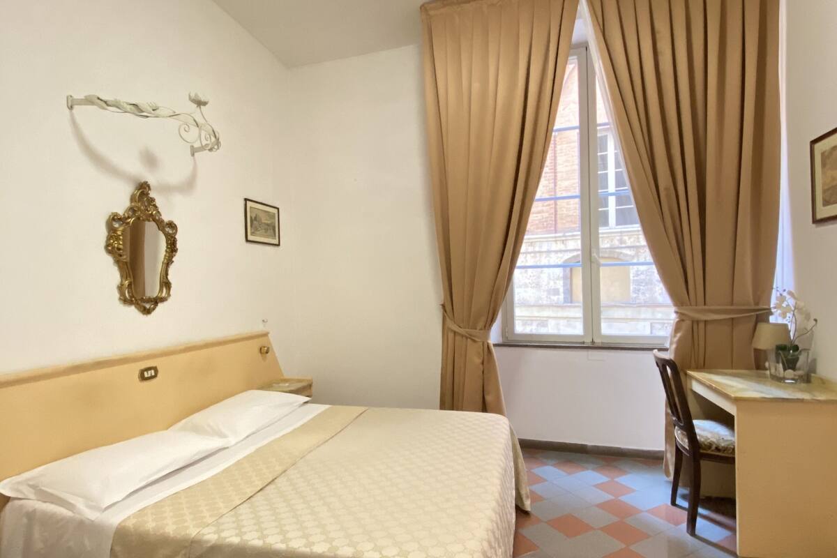 A hotel room with a bed, a desk, a chair, a window with curtains, a wall-mounted mirror, and a picture.