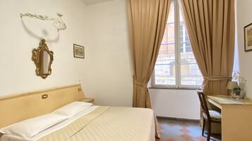 A hotel room with a bed, a desk, a chair, a window with curtains, a wall-mounted mirror, and a picture.