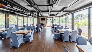 A restaurant with wooden tables and grey chairs, large windows with a view of greenery and buildings, and visible ceiling pipes.