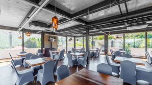 En modern restaurang med högt i tak, synliga ventilationskanaler och stora fönster som ger utsikt över grönområden. Bord och stolar är placerade för att skapa en trevlig matplats.