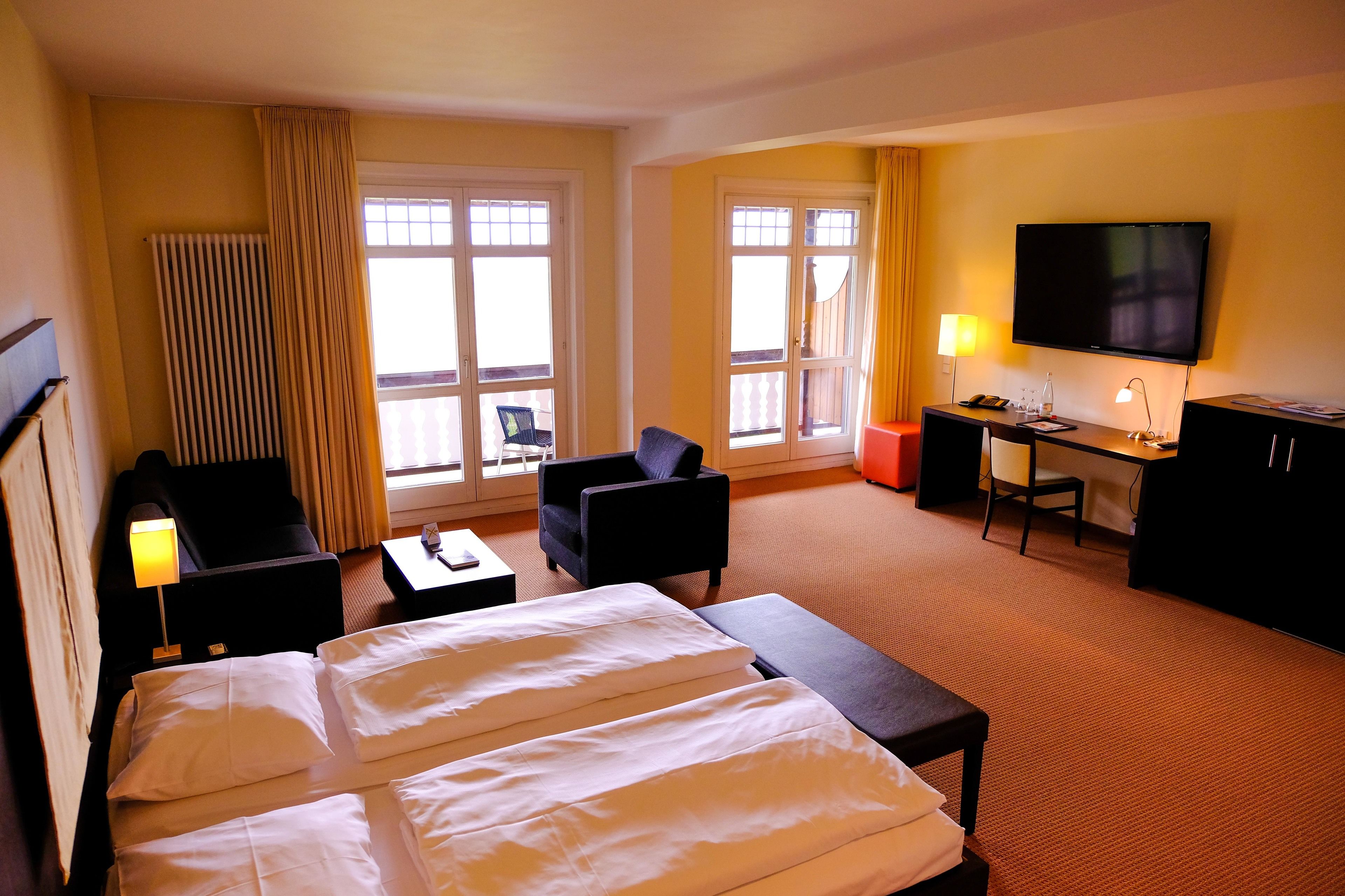 A hotel room with a bed, two armchairs, a desk, a television, and a window with curtains.