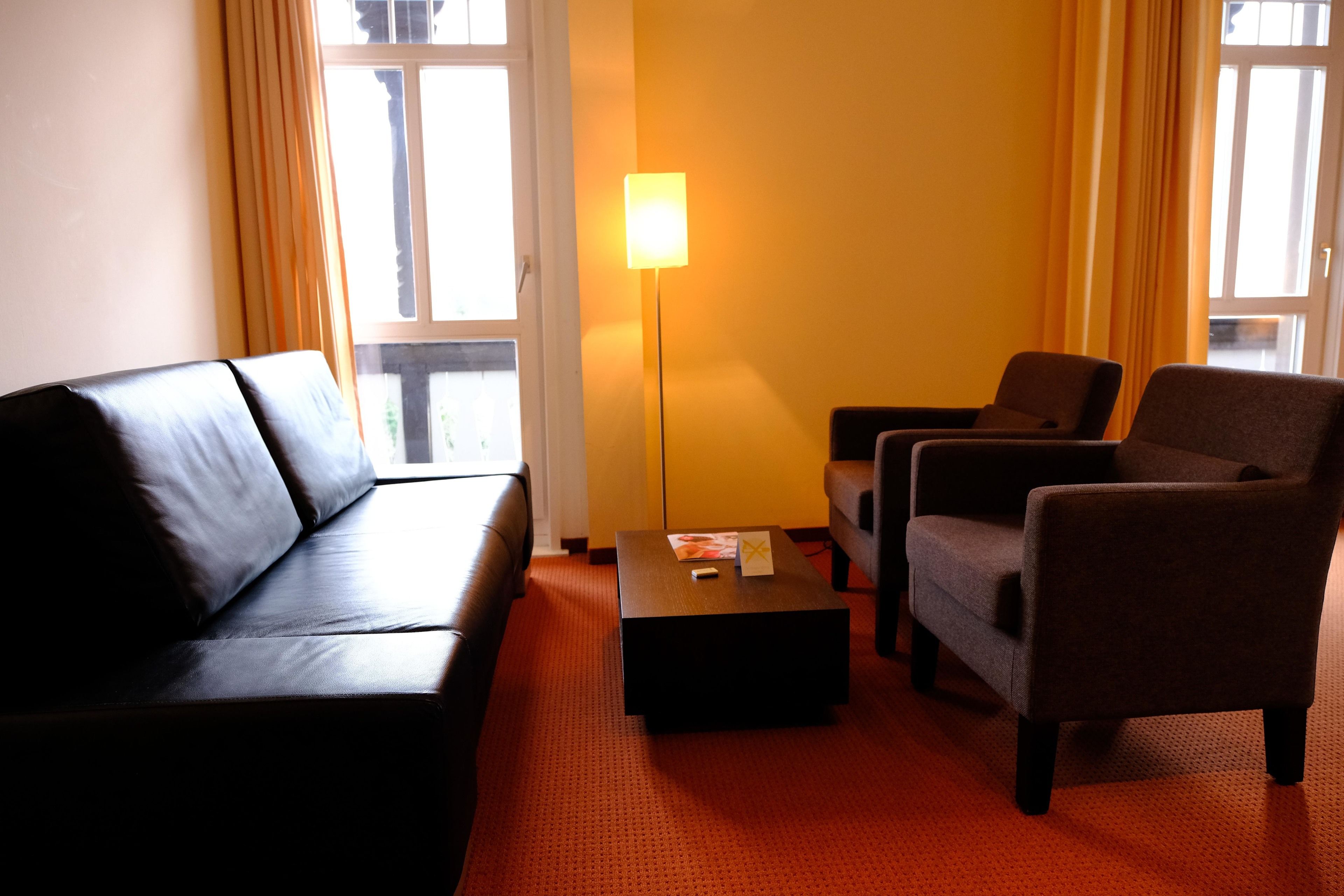 A hotel room with a black leather sofa, two armchairs, a small table, and a floor lamp.