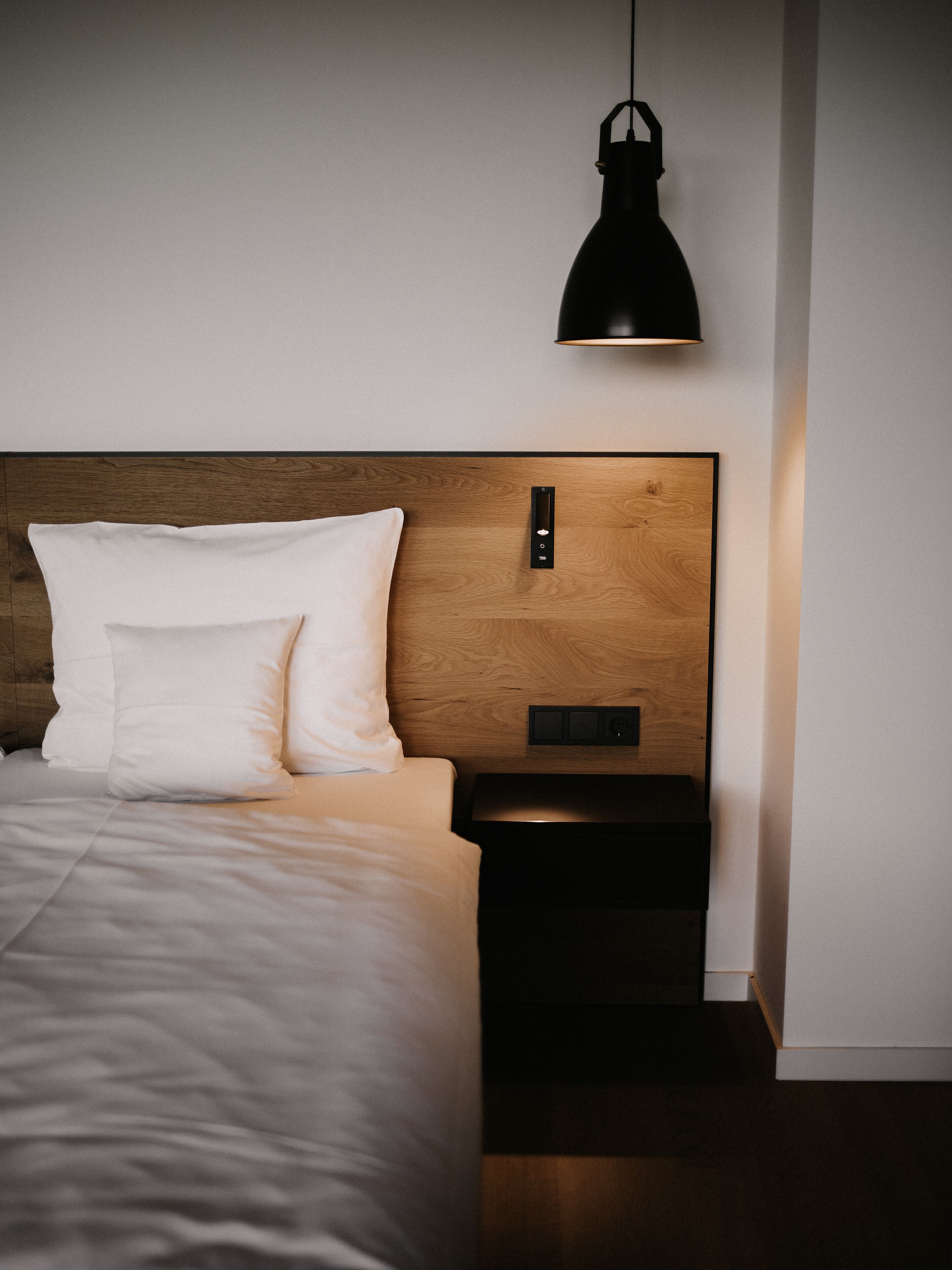 A neatly made bed with white linens and pillows, a wooden headboard, a black bedside table with a switch, and a pendant light above.