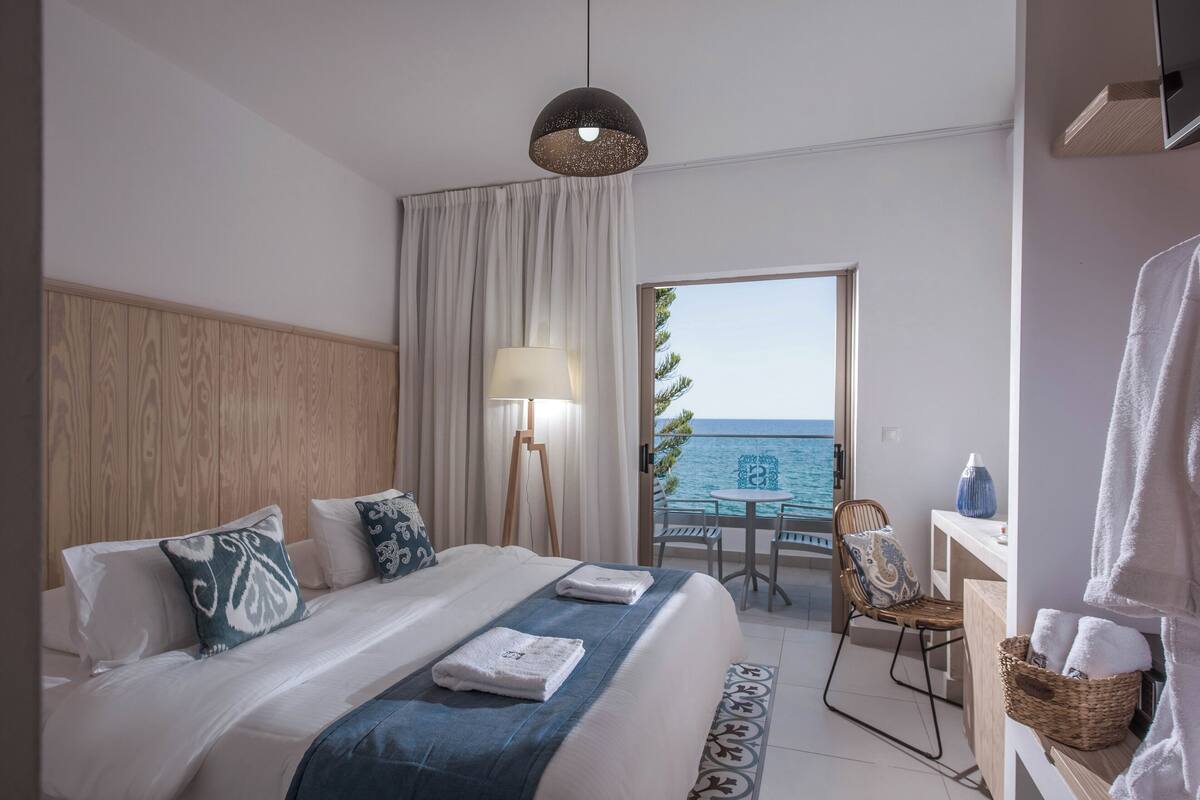 A hotel room with a bed, a desk, a chair, a lamp, and a view of the sea.
