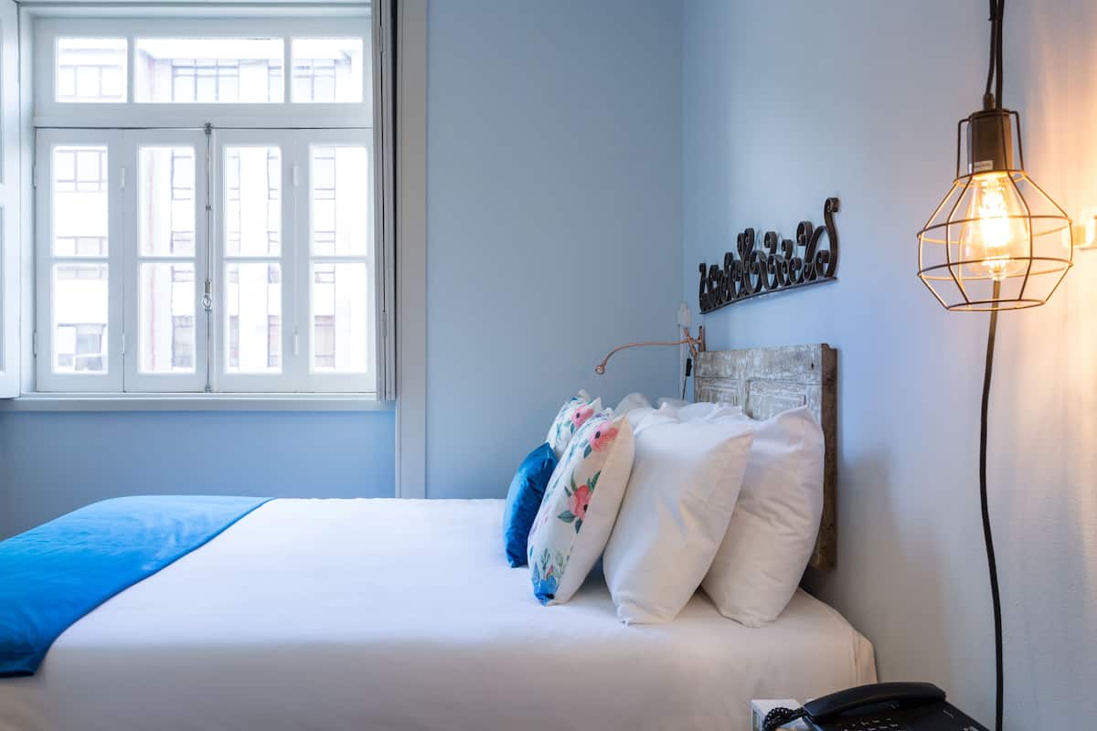 Un dormitorio con cama, almohadas, una mesita de noche, una lámpara y una ventana.