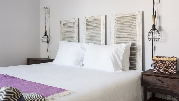 A neatly made bed with white linens, a purple throw blanket, and a striped hat on the side. There are two bedside tables with lamps and a woven basket on one. - The Bohemian Place (Aveiro)