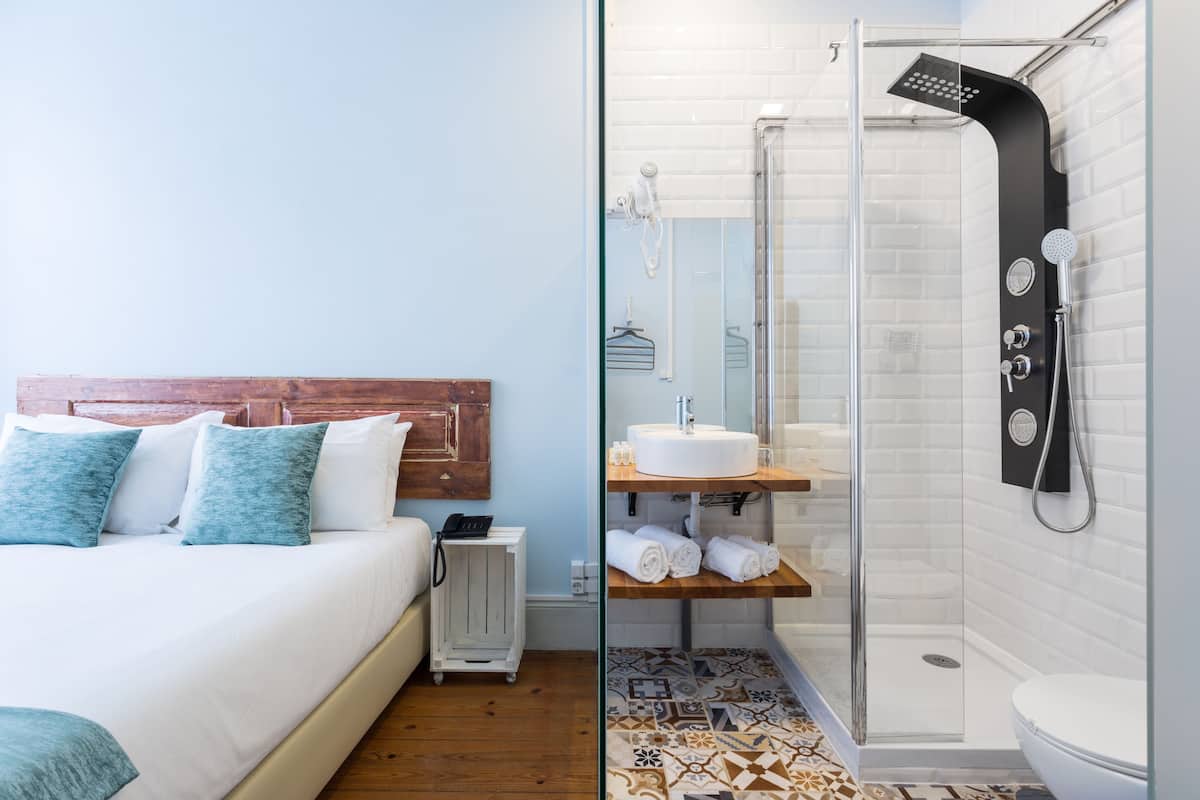Una habitación de hotel con una cama, cabecera de madera, una mesita de noche con un teléfono y un armario pequeño. El baño tiene una cabina de ducha de cristal, un lavabo blanco y un inodoro.