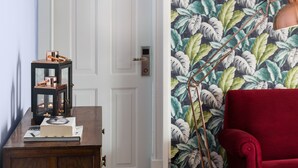 A room with a wooden desk, a red chair, a door with a modern handle, and a wall with a tropical leaf pattern. - The Bohemian Place (Aveiro)