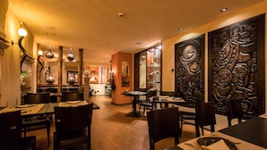 Restaurante com mesas e cadeiras de madeira, área de bar e painéis decorativos na parede.