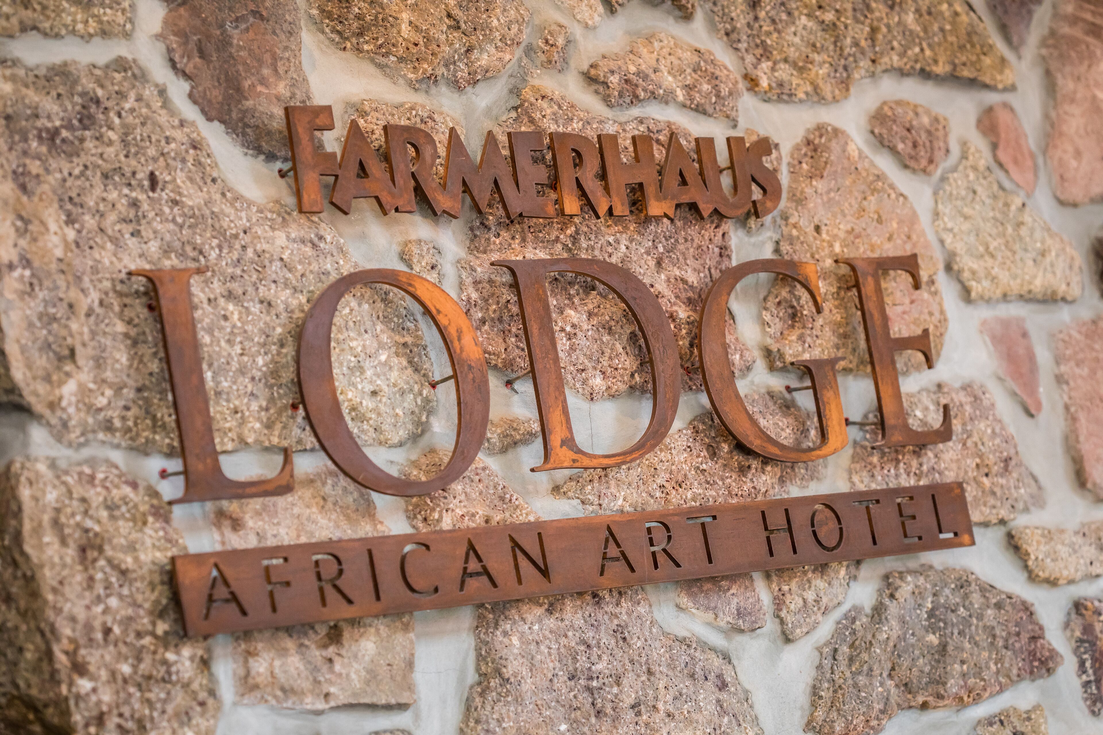 A sign for the Farmerhals Lodge, an African Art Hotel, with a rustic stone wall background.