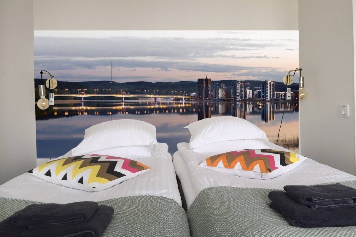 A room with two beds facing a cityscape and a body of water.