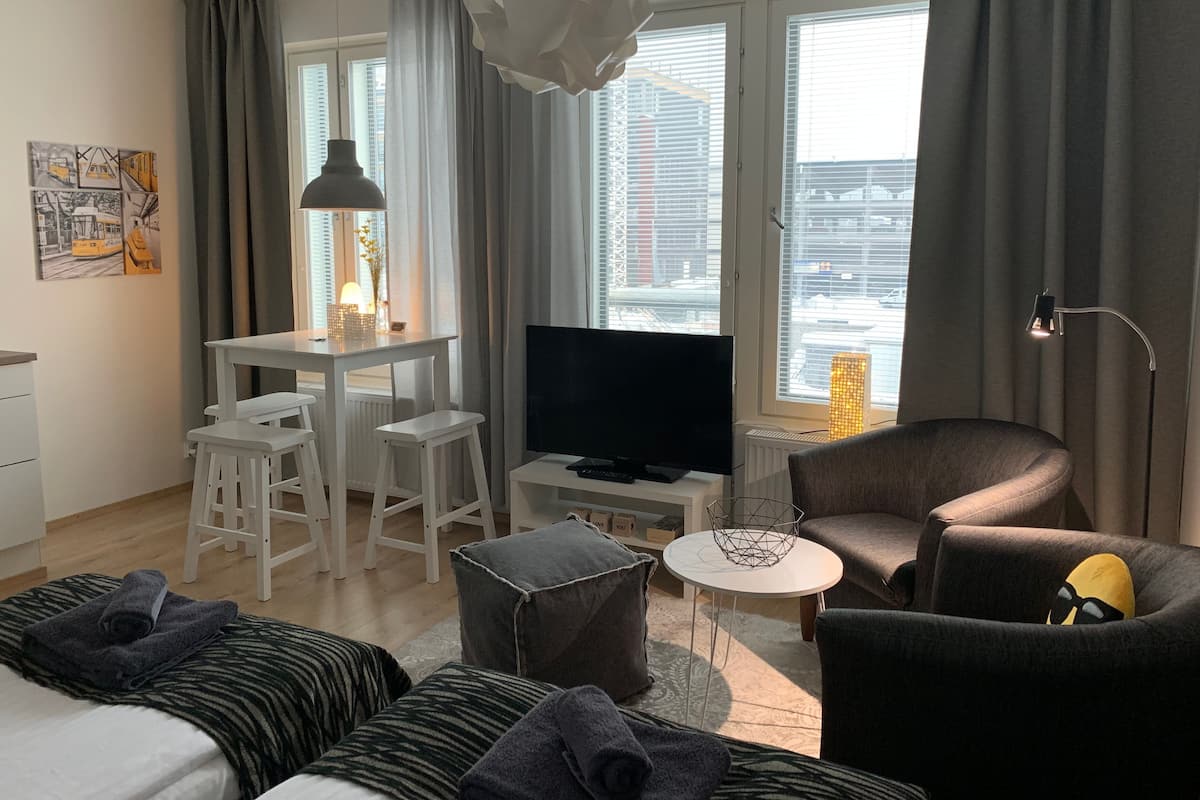 A modern hotel room with a bed, a desk with a TV, a chair, and a view of the city through large windows.