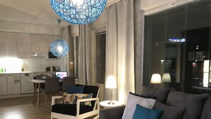 Un salon moderne avec un canapé gris, un fauteuil à bascule blanc et un coin repas comprenant une table et des chaises. Au fond, on aperçoit une cuisine avec des meubles blancs et une table à manger.