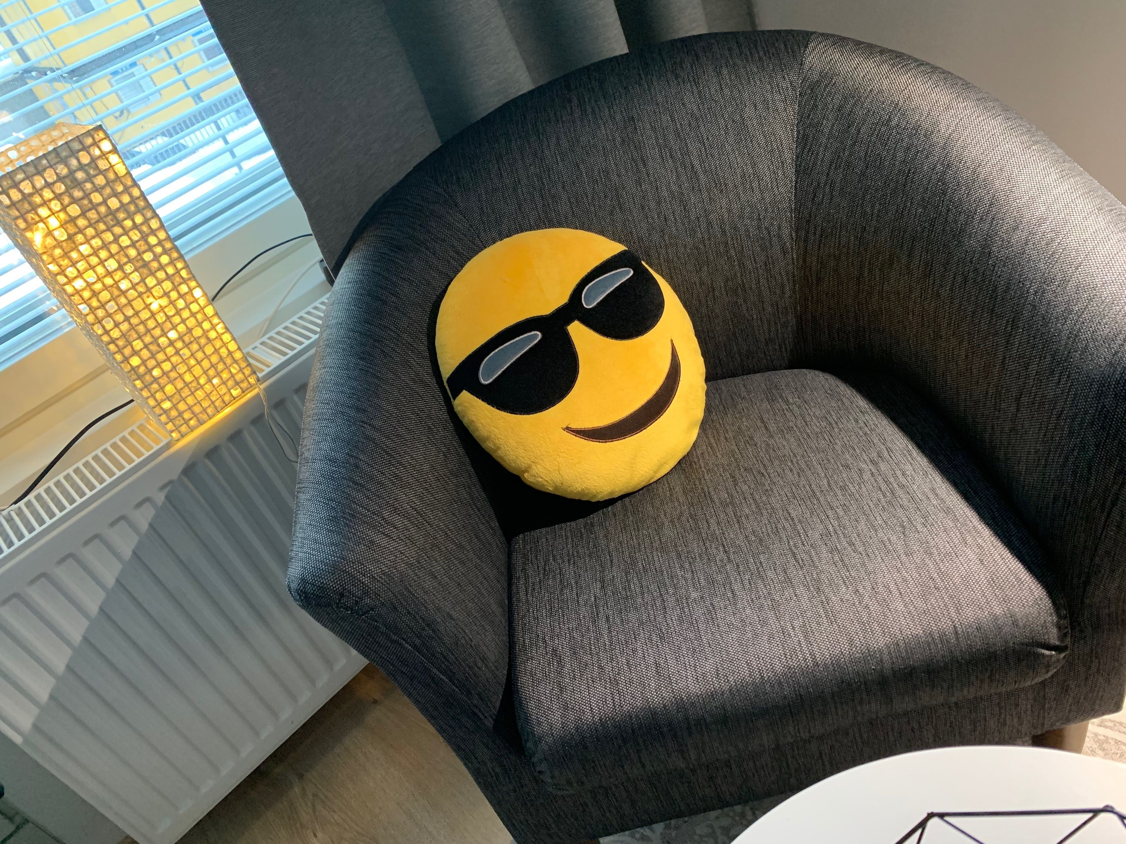 A grey armchair with a yellow pillow featuring a smiling face and sunglasses.